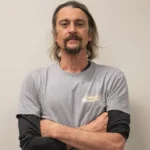 Casey with Heavenly Garage Doors poses with arms crossed in a gray tee against a gray wall.