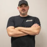 Tim Carnes with Heavenly Garage Doors poses with arms crossed in a black hat and black tee against a gray wall.